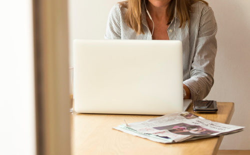 Woman at desk on laptop with newspaper and phone to the side